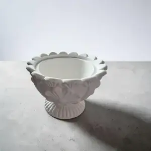 Coupe décorative blanche sur pied, avec motifs floraux sculptés en relief et bordure festonnée, photographiée sur fond gris clair.