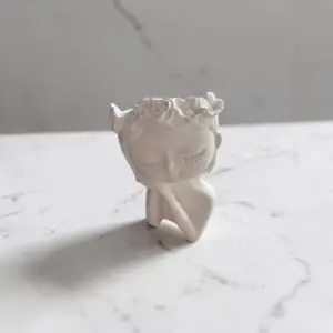 Objet décoratif sculpté représentant un visage féminin aux yeux fermés, avec une couronne de fleurs et une main posée sous le menton.