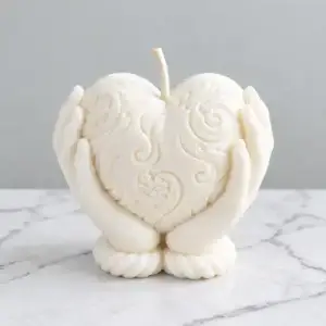 Bougie cœur décorative blanche, ornée de motifs floraux et de volutes en relief, soutenue par deux mains sculptées, sur fond blanc effet marbre.